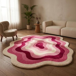 Puede incluir: Una alfombra abstracta ondulada en tonos rosas y blancos. Presenta patrones concéntricos en capas de varios tonos de rosa, desde un centro magenta oscuro hasta un borde blanco roto suave. Se encuentra sobre un suelo de madera en un salón.