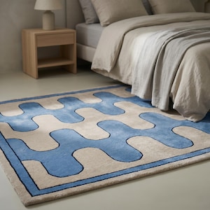 Peut inclure: Un tapis rectangulaire présentant un motif ondulé bleu répétitif sur un fond beige. Le tapis est doté d'une bordure bleue unie qui encadre le design sinueux, lequel s'étend sur toute la surface dans un motif de vagues interconnectées.