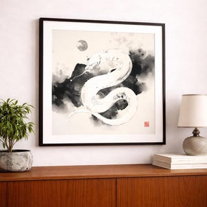 以下が含まれることがあります： 黒と白の墨絵風の蛇が描かれた正方形の額入りウォールアート。暗く煙るような背景に小さな月が浮かび、プリントの右下隅には赤い正方形の印が押されている。