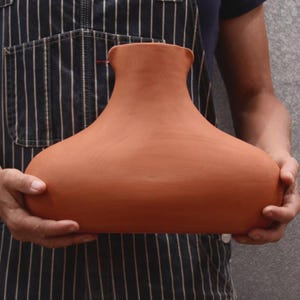 May include: A wide, terracotta-colored ceramic vase with a smooth, matte finish. The vessel features a broad, rounded base that tapers upward into a narrow, open neck with a subtle spout-like indentation at the top rim, showcasing a simple form.