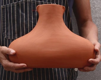 Peut inclure: Un large récipient en argile de couleur terre cuite avec une finition lisse et mate. L'objet présente une base large et arrondie qui se rétrécit vers le haut en un col étroit et ouvert avec un bord légèrement évasé, offrant une silhouette symétrique.