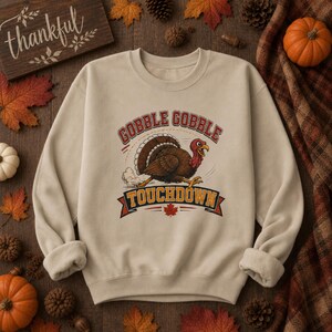 May include: A beige crewneck sweatshirt featuring a graphic of a running turkey in the center. The text above the turkey reads GOBBLE GOBBLE and the text below on a banner reads TOUCHDOWN. A small red maple leaf is centered below the turkey.
