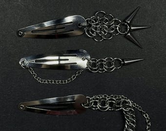 May include: A set of five silver-toned metal hair clips featuring chainmail links, spikes, a star charm, and delicate chains. The clips are arranged vertically, showcasing various designs including single spikes, a star, and linked chains.