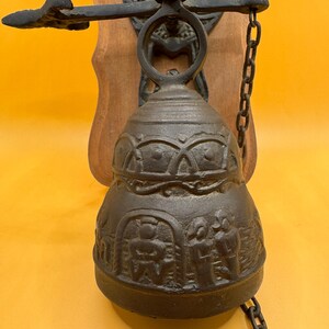May include: A dark brown cast iron wall-mounted bell featuring a relief design of figures and patterns. The bell is attached to a wooden backplate by a metal bracket and includes a hanging metal chain for ringing, showcasing a textured finish.