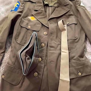 May include: An olive drab military-style jacket featuring brass buttons, four front pockets, and shoulder epaulets. A blue and yellow patch is on the right shoulder, and a beige necktie is draped over the front alongside a folded garrison cap.