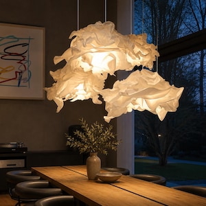 May include: Three white, cloud-like pendant lights hang above a wooden dining table. The lights have a textured, ruffled appearance, casting a warm glow. A vase with greenery and a bowl are on the table. The room has a modern, minimalist design.