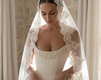 May include: A sheer white bridal veil featuring intricate floral lace trim with pearl embellishments, draped over the head. The veil is paired with a cream-colored corset-style bodice dress that has a gathered neckline and a structured silhouette.