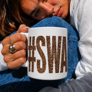 May include: A white ceramic mug featuring the text #SWA printed in a bold, block font with a brown and black leopard print pattern. The mug has a smooth, cylindrical body and a curved handle on the side, designed for holding hot or cold beverages.