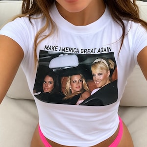 May include: A white short-sleeved t-shirt featuring a rectangular graphic print of three women sitting in a car. Above the image, the text reads "MAKE AMERICA GREAT AGAIN" in black capital letters. The shirt has a crew neckline and a fitted silhouette.