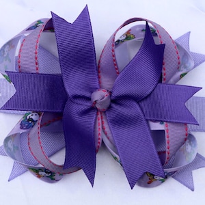 May include: A layered hair bow constructed from purple grosgrain ribbon and a lighter lavender ribbon featuring a repeating cartoon character print. The design includes a central knot and multiple pointed ribbon tails extending outward.