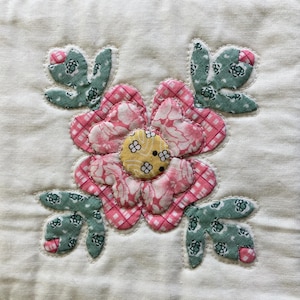 May include: A square fabric quilt block featuring a central appliqued flower with pink checkered petals and a yellow center. Four green floral patterned leaves with small pink accents surround the flower, all set against a white background.