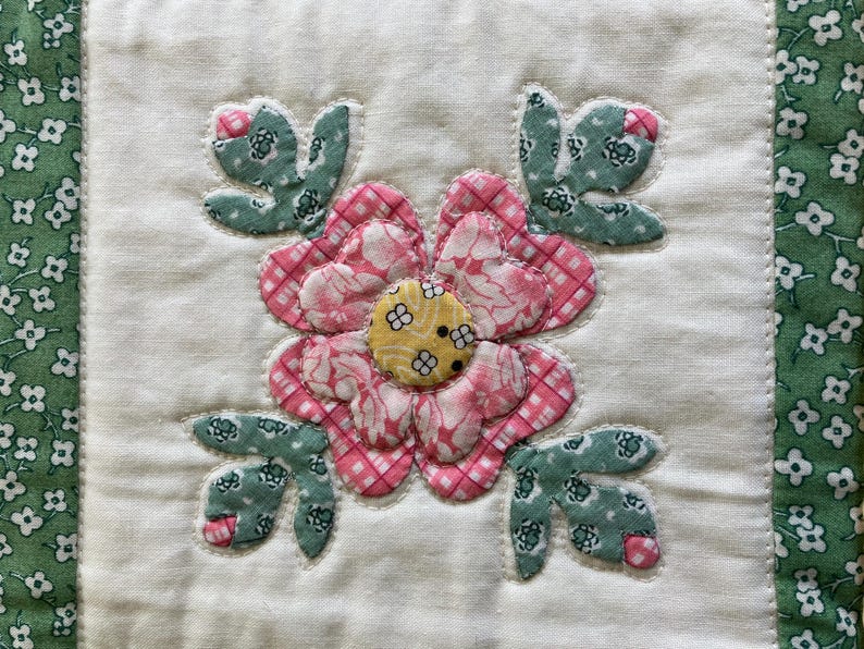 May include: A square fabric quilt block featuring a central appliqued flower with pink checkered petals and a yellow center. Four green floral patterned leaves with small pink accents surround the flower, all set against a white background.