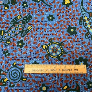 Può includere: Un tessuto blu con un motivo ripetuto di lucertole e tartarughe verde scuro con accenti geometrici gialli e arancioni, intervallati da fiori verde scuro. Un righello di legno mostra una scala di 15 cm nella parte inferiore della stampa.