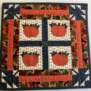 May include: A square fabric quilt featuring four central blocks with orange pumpkin appliqués on a cream background with acorn patterns. The quilt is bordered by dark blue gingham fabric and a wide band of orange and brown autumn leaf prints.