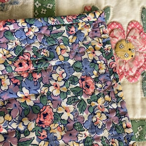 May include: A quilted fabric piece featuring a dense floral pattern in shades of blue, pink, yellow, and green. The fabric is stitched with a leaf-shaped quilting design. To the right, a separate cream-colored fabric block displays a large pink and green floral appliqu&eacute; with a yellow center.
