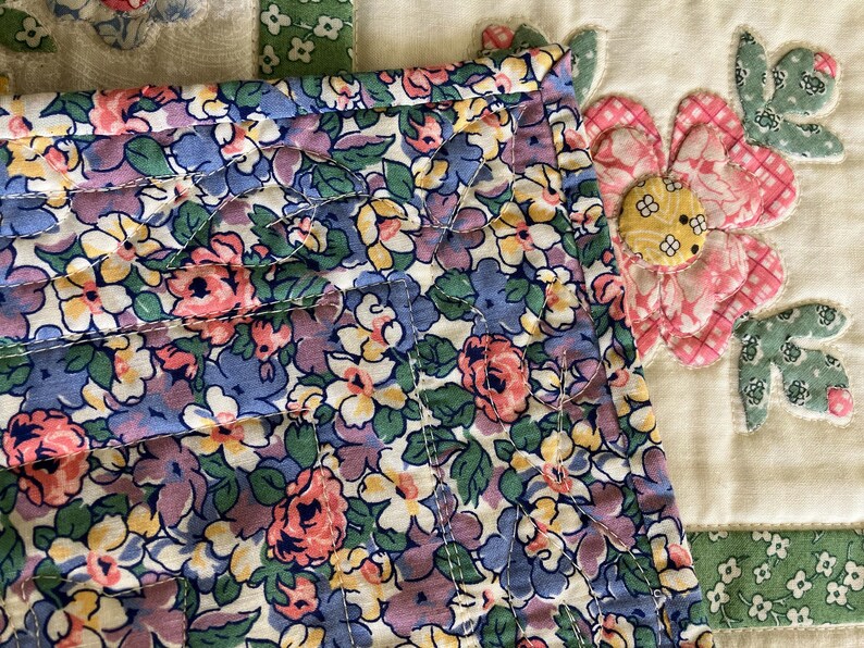 May include: A quilted fabric piece featuring a dense floral pattern in shades of blue, pink, yellow, and green. The fabric is stitched with a leaf-shaped quilting design. To the right, a separate cream-colored fabric block displays a large pink and green floral appliqu&eacute; with a yellow center.