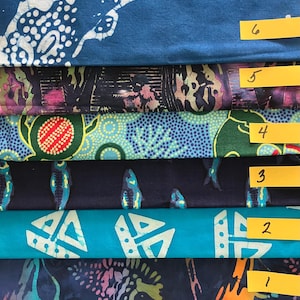 Könnte beinhalten: Sechs gestapelte Stoffmuster mit gelben Papieretiketten, die von eins bis sechs nummeriert sind. Die Muster umfassen blau-weißes Batik, Dunkellila mit abstrakten Formen, Türkis mit Korallenmotiven, Dunkelblau mit Fischen und Türkis mit weißen geometrischen Formen.