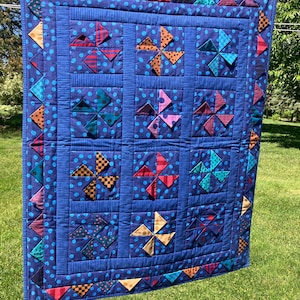 May include: A rectangular quilt featuring a dark blue fabric with light blue polka dots. The quilt is composed of nine square blocks, each containing a multi-colored pinwheel pattern. The border is decorated with small, colorful fabric triangles.