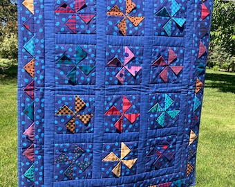 May include: A rectangular quilt featuring a dark blue fabric with light blue polka dots. The quilt is composed of nine square blocks, each containing a multi-colored pinwheel pattern. The border is decorated with small, colorful fabric triangles.