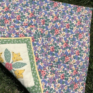 May include: A quilted fabric piece featuring a dense floral pattern with pink, purple, and yellow blossoms on a green leafy background. One corner is folded back to reveal a white square panel with a green border and a central floral applique.
