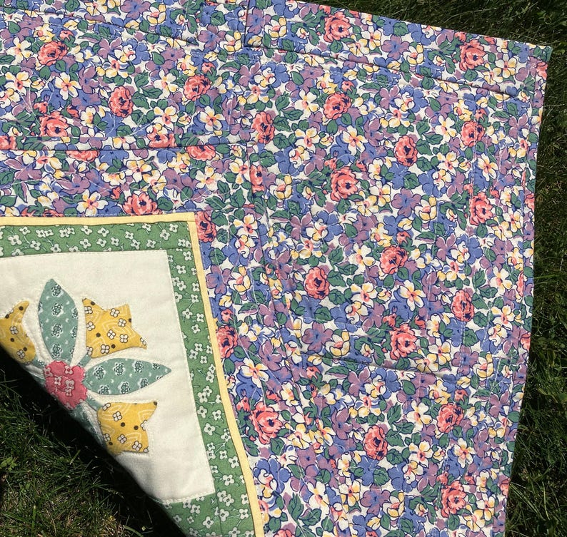 May include: A quilted fabric piece featuring a dense floral pattern with pink, purple, and yellow blossoms on a green leafy background. One corner is folded back to reveal a white square panel with a green border and a central floral applique.