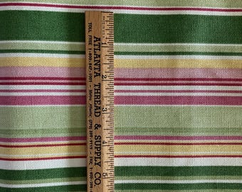 May include: A woven fabric featuring a repeating horizontal striped pattern in shades of dark green, light green, yellow, pink, and white. A wooden ruler is placed vertically on the fabric, showing a measurement scale of up to 6 inches.