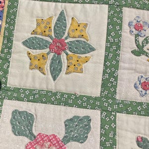 May include: A patchwork quilt featuring square blocks with floral applique designs. The fabric includes white squares with yellow, pink, and green floral patterns, separated by green fabric strips with a small white flower print. Visible stitching outlines each applique shape.
