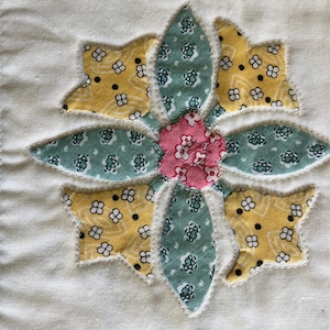 May include: A fabric quilt block featuring a central flower appliqu&eacute; made of yellow, teal, and pink patterned cotton pieces. The flower is stitched onto a solid off-white background, bordered by a green fabric strip with a white floral print.