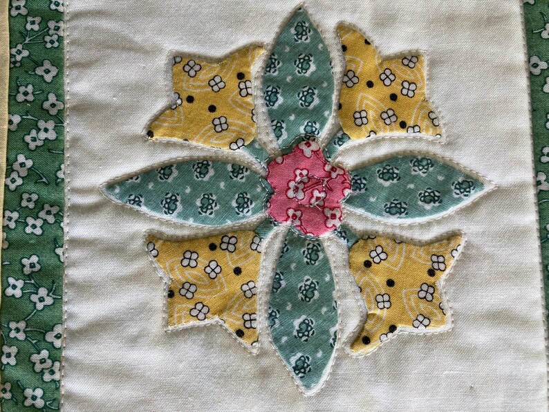 May include: A fabric quilt block featuring a central flower appliqu&eacute; made of yellow, teal, and pink patterned cotton pieces. The flower is stitched onto a solid off-white background, bordered by a green fabric strip with a white floral print.