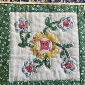 May include: A quilted fabric panel featuring a central yellow sun-shaped applique with a pink floral square center, surrounded by four light blue and pink floral blossoms on green stems, all set against a white background with green floral borders.