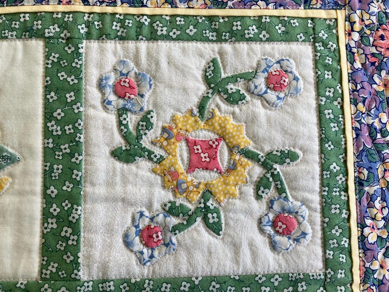 May include: A quilted fabric panel featuring a central yellow sun-shaped applique with a pink floral square center, surrounded by four light blue and pink floral blossoms on green stems, all set against a white background with green floral borders.