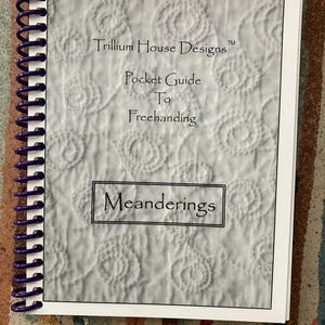 May include: A spiral-bound instructional booklet titled Trillium House Designs Pocket Guide To Freehanding Meanderings. The cover features a white background with a subtle, repeating circular quilted pattern and dark purple plastic binding coils.