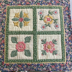 May include: A square quilted wall hanging featuring four distinct floral applique blocks on a cream background. Each block displays different fabric flowers in shades of pink, yellow, and green, framed by a green floral border and a floral outer edge.