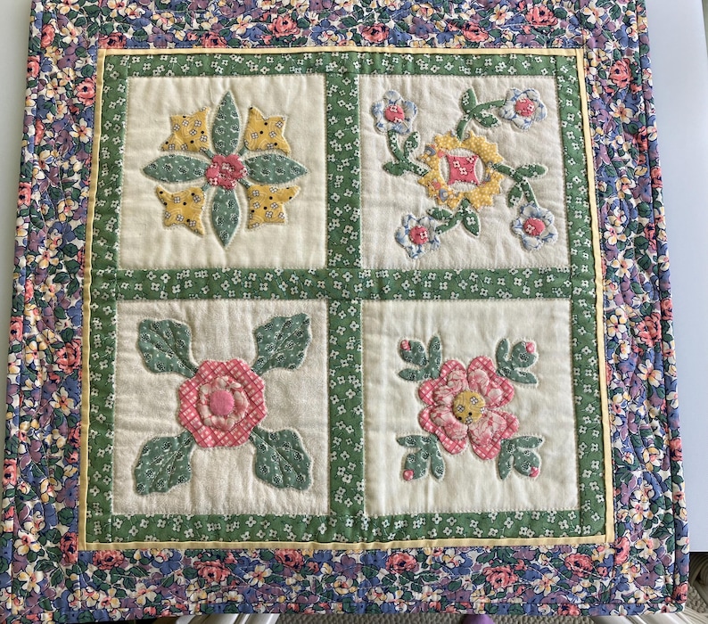 May include: A square quilted wall hanging featuring four distinct floral applique blocks on a cream background. Each block displays different fabric flowers in shades of pink, yellow, and green, framed by a green floral border and a floral outer edge.