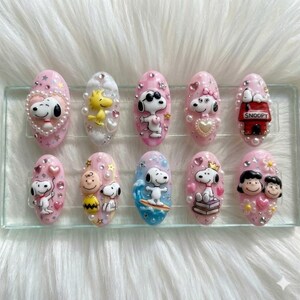 May include: A set of ten oval-shaped press-on nails featuring Peanuts characters. Each nail is decorated with 3D figures of Snoopy, Charlie Brown, and Lucy, accented with pearls, rhinestones, stars, hearts, and flowers on pink and blue backgrounds.