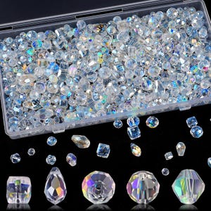 May include: A clear plastic rectangular storage container filled with a large assortment of multifaceted, iridescent glass beads in various shapes and sizes, including round, teardrop, and cube forms, with several loose beads scattered below.