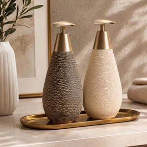 May include: Two teardrop-shaped soap dispensers with textured surfaces, one in dark gray and one in cream, featuring gold-toned pump mechanisms. They are arranged side-by-side on an elongated, matching gold-toned metal tray for a coordinated look.