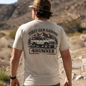 May include: A beige short-sleeve t-shirt featuring a graphic on the back of a white SUV, pine trees, and mountains. The text reads FIRST GEN GARAGE, 4RUNNER, and 1984-1989. The design includes orange and yellow horizontal stripes below the vehicle.