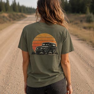 May include: An olive green short-sleeve t-shirt featuring a graphic on the back. The design displays a black off-road vehicle parked in front of a large, orange, horizontally striped circular sun, with small silhouettes of pine trees at the base.