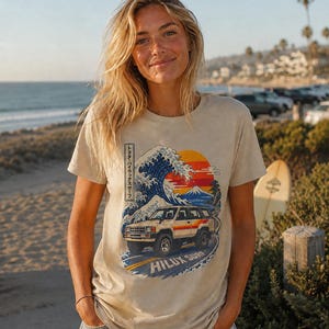May include: A beige short-sleeve t-shirt featuring a graphic print of a Toyota Hilux Surf vehicle driving on a road. The design includes a large blue wave, a sunset, Mount Fuji, and Japanese text. The text HILUX SURF is printed at the bottom.