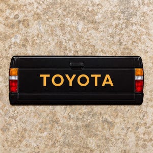 May include: A black Toyota truck tailgate featuring the word TOYOTA in bold yellow lettering centered on the panel. The tailgate includes a central black handle and rectangular red, white, and amber taillight assemblies on both the left and right sides.