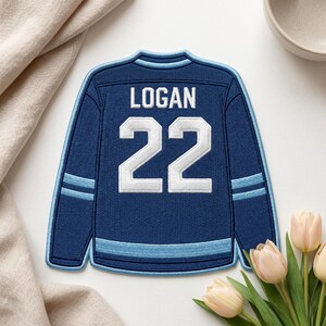 Puede incluir: Un posavasos con forma de camiseta de hockey azul marino con el nombre "LOGAN" y el número "22" en blanco. El posavasos tiene detalles en azul claro en las mangas y en la parte inferior. El posavasos está sobre una superficie blanca.