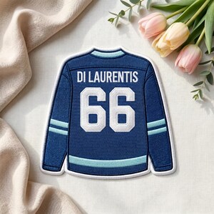 Puede incluir: Un parche de camiseta de hockey azul marino con el nombre "DI LAURENTIS" y el número "66" en blanco. La camiseta tiene rayas azul claro en las mangas y en la parte inferior. El parche está sobre una superficie color crema con tulipanes rosas.
