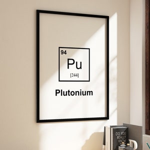 May include: A black framed poster with a white background and black text. The poster features the chemical element Plutonium, with the atomic number 94 and atomic weight 244. The poster is hanging on a white wall in a home office setting. A wooden desk with a lamp, books, and other office supplies is in the foreground.