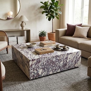 May include: A large, square, low-profile coffee table crafted from white marble with prominent, deep purple veining. The table surface holds a small white bowl, a stack of two books, a set of keys, and a dark circular tray with two ceramic mugs.