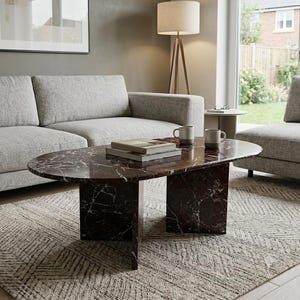 May include: An oval coffee table made of dark red marble with white veining, featuring two thick slab legs. Two stacked books and two ceramic mugs are placed on the smooth surface of the table, which is positioned in a living room setting.
