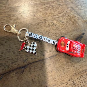 Op de afbeelding: Een gepersonaliseerde sleutelhanger met de naam "BROOKS" gespeld met witte letterkralen. De sleutelhanger bevat een rode raceauto-bedel, een hartvormige bedel met een dambordpatroon en een bliksembedel. Een goudkleurige sluiting is bevestigd.