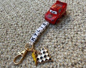 May include: A keychain featuring a red Lightning McQueen toy car attached to a string of white beads spelling the name KYLIE. The keychain includes a gold-toned lobster clasp, a yellow lightning bolt charm, and a black and white checkered heart charm.