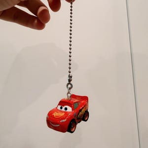 May include: A red Lightning McQueen toy car keychain featuring the number 95 on the side, Rust-eze branding on the hood, and large blue eyes on the windshield. The miniature vehicle is attached to a silver-toned metal ball chain connector.
