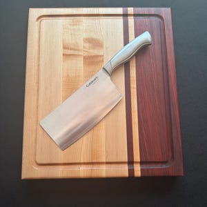 May include: A rectangular wooden chopping board featuring light maple and dark mahogany wood strips with a juice groove, topped with a stainless steel Cuisinart cleaver. The knife has a metallic handle and a wide, flat blade with the brand name engraved.